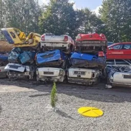 Stos pogniecionych i zniszczonych karoserii samochodowych na złomowisku, z widocznymi elementami różnych marek i kolorów, ułożonych w nieładzie na tle zieleni i szarego żwiru.