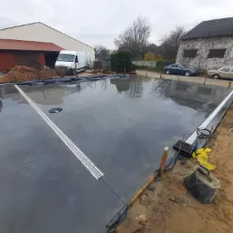 Świeżo wylana, gładka, szara powierzchnia z wbudowanymi liniowymi odpływami, przygotowana pod posadzkę przemysłową, widoczne narzędzia i drewniane szalunki, w tle budynki i zaparkowane samochody.