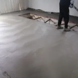 Wyrównywanie wylewki betonowej za pomocą zacieraczki mechanicznej, widoczne świeże ślady butów na powierzchni oraz niezabezpieczony otwór w podłodze.