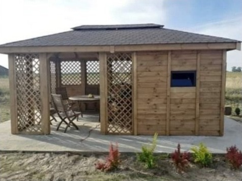 Drewniana altana z kratownicowymi ścianami i ciemnym dachem, wyposażona w okrągły stół i krzesła wewnątrz, usytuowana na betonowej płycie w ogrodzie.