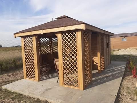 Gotowa, drewniana altana ogrodowa z brązowym dachem, kratowanymi ścianami i betonową podstawą, widok z zewnątrz w słoneczny dzień.