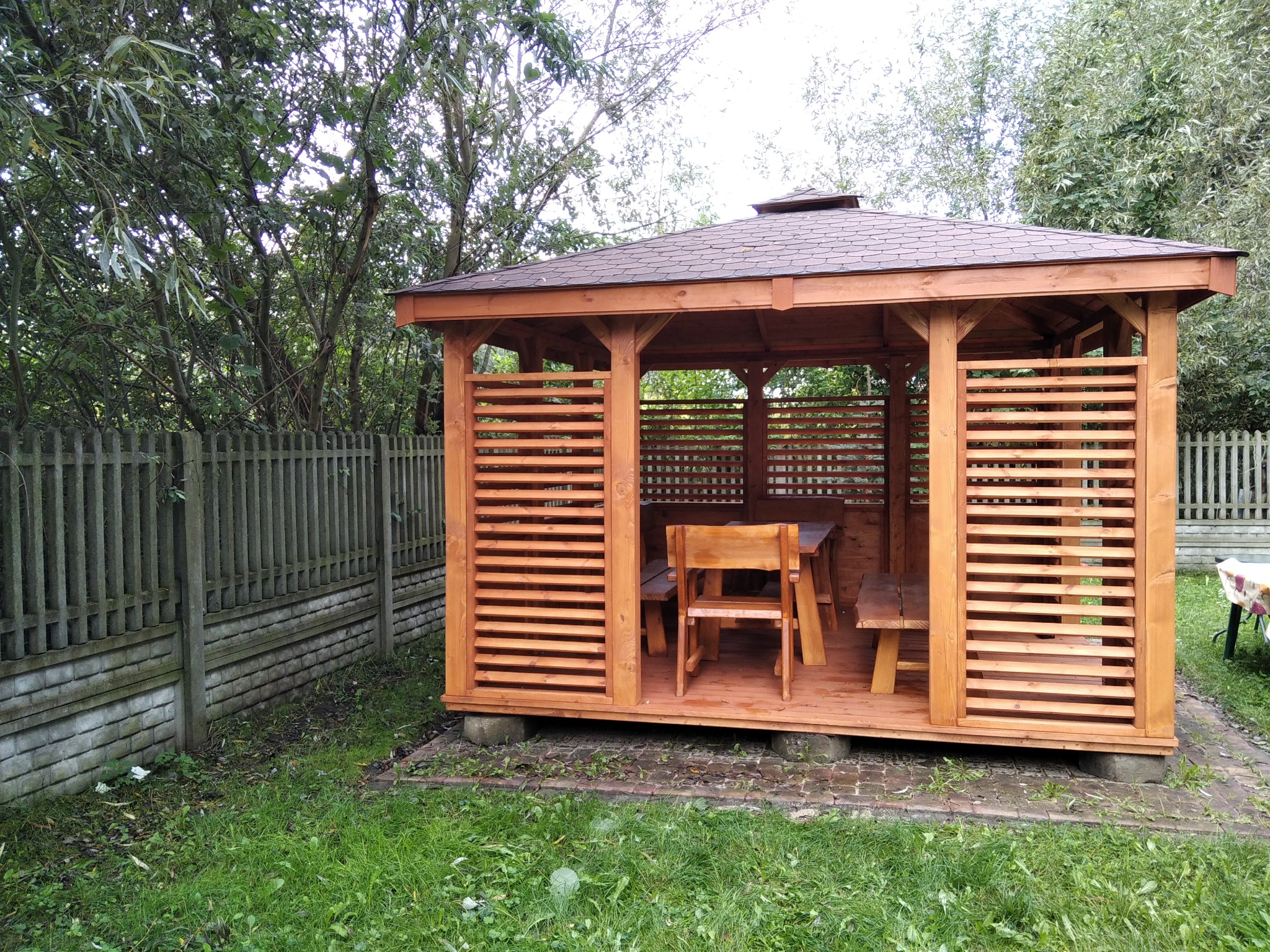 Drewniana altana z brązowym dachem i poziomymi listwami, na betonowych podstawach, z drewnianym stołem i krzesłami w środku, otoczona zielenią.