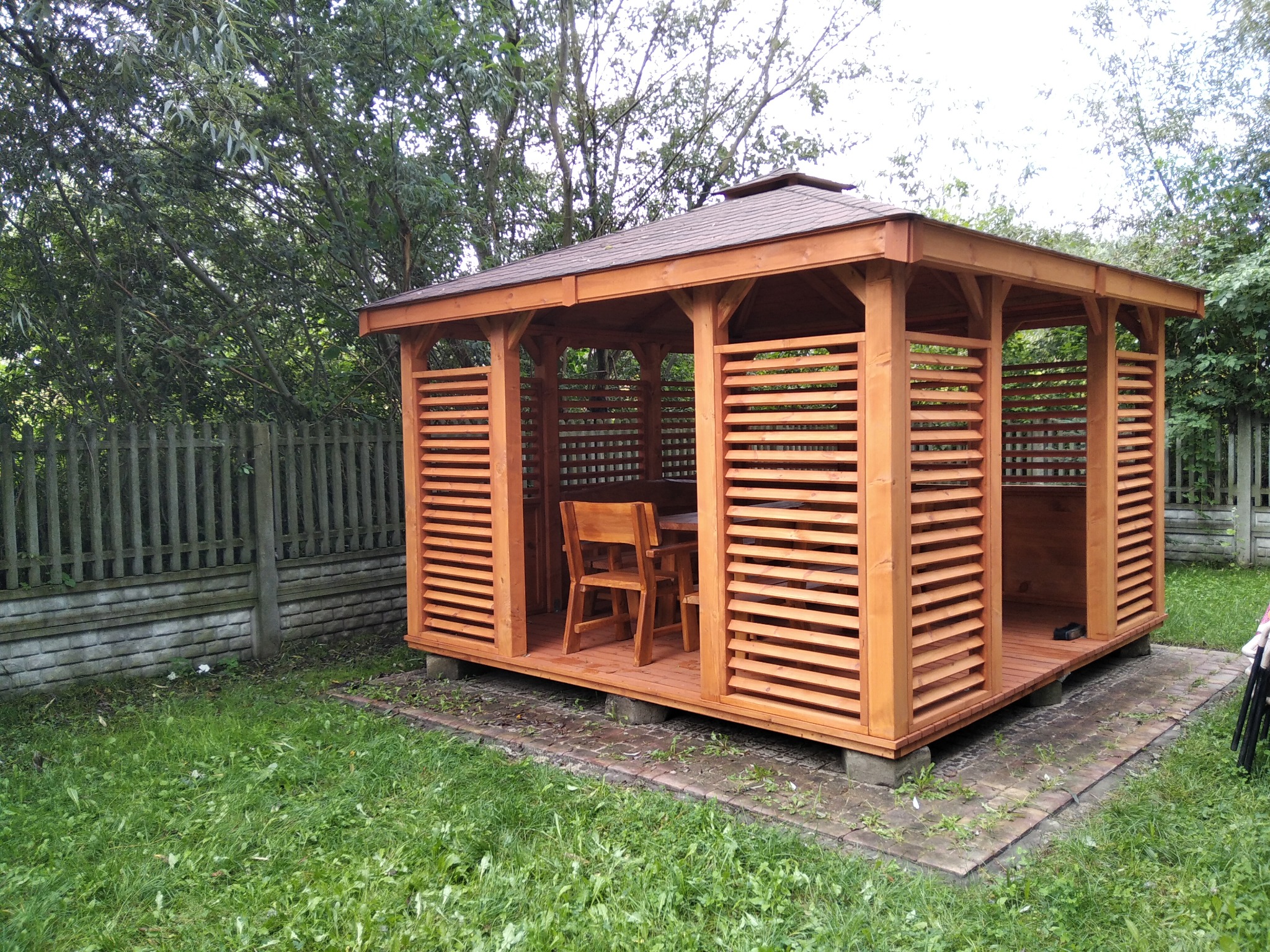 Drewniana altana z poziomymi listwami, widok z zewnątrz na stół i krzesła w środku, otoczona zielenią i drewnianym płotem.