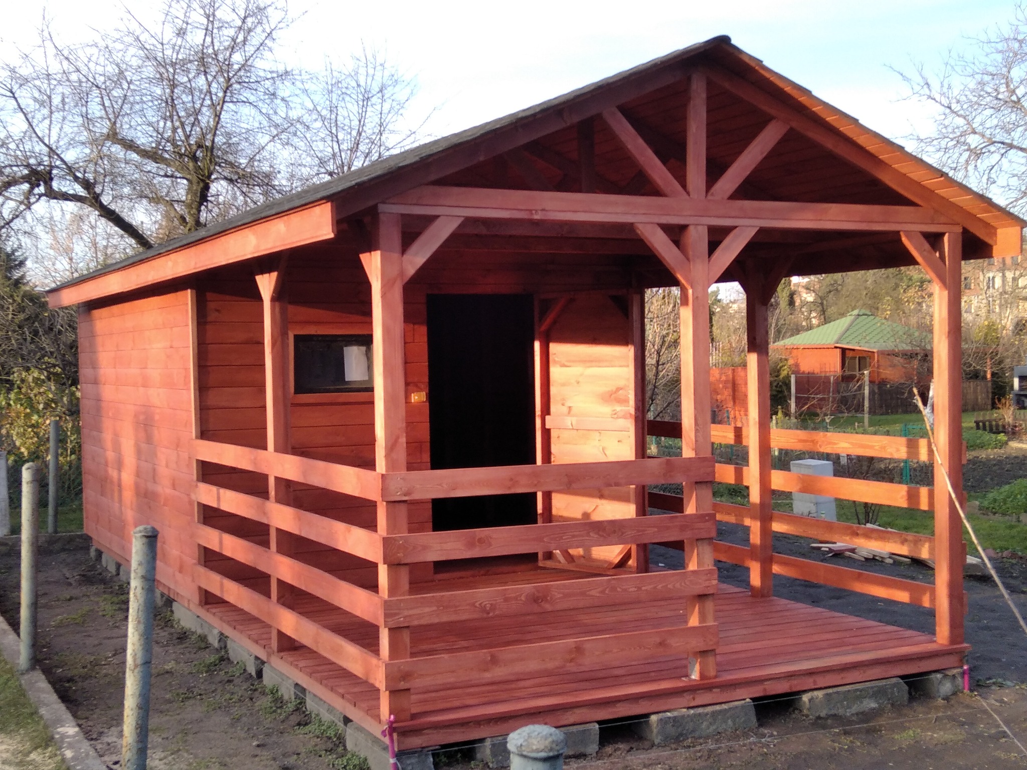 Czerwona, drewniana konstrukcja z zadaszeniem i otwartym tarasem, w tle widoczna inna budowla i drzewa bez liści.