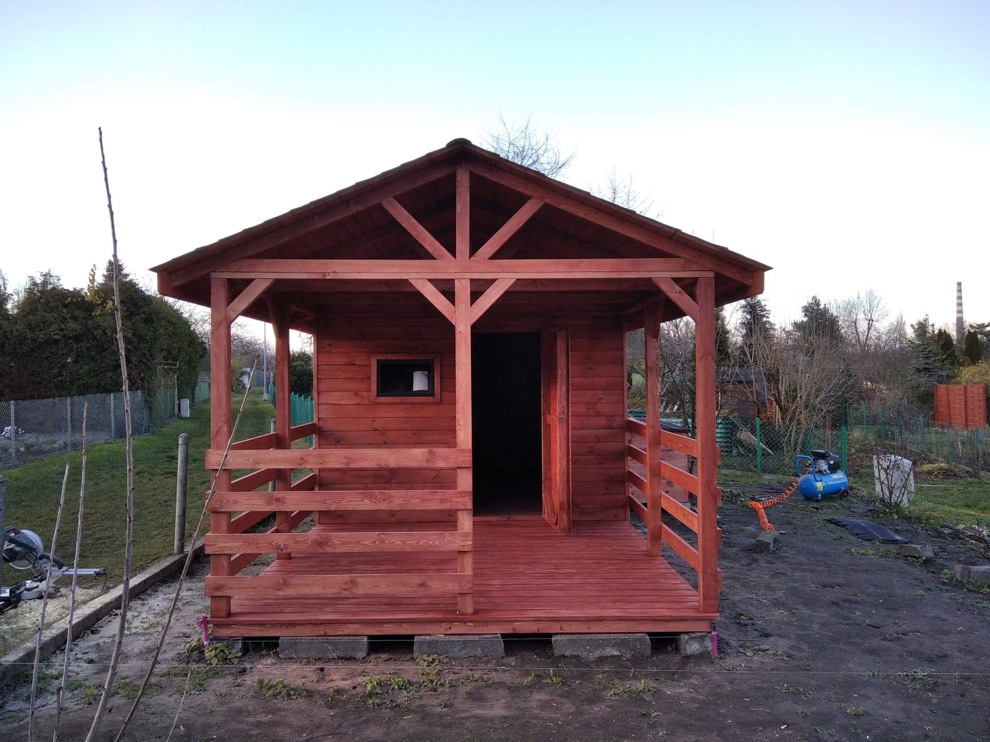 Nowa, drewniana altana w kolorze czerwono-brązowym, z widoczną konstrukcją dachu i balustradami, umieszczona na betonowych bloczkach w ogrodzie, obok leży niebieski kompresor.