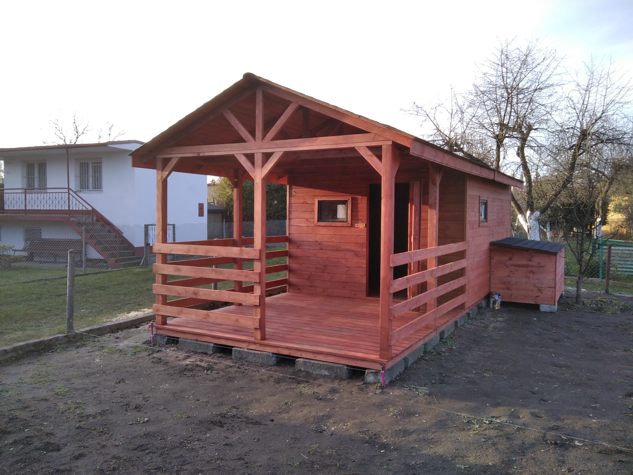 Nowo wybudowana, pomalowana na czerwono altana z drewnianą podłogą, umieszczona na betonowych bloczkach, z dodatkową zabudową z tyłu, na tle domu i drzew bez liści.