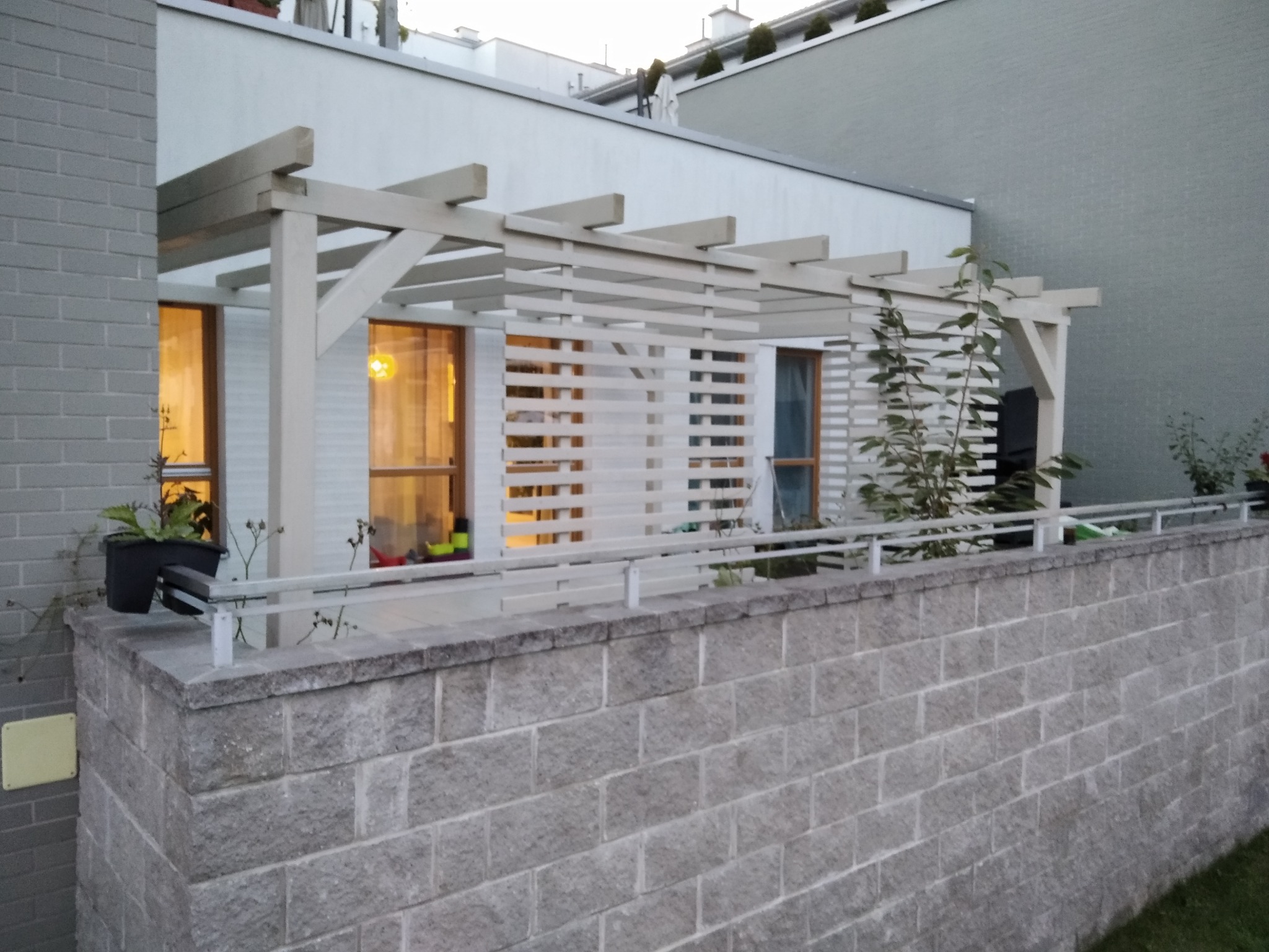 Kremowa pergola z poziomymi listwami przysłaniająca okna domu, widoczna zza murku z szarej kostki, z donicami na górze.