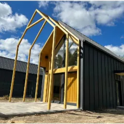 Nowoczesny dom z czarną elewacją i drewnianą konstrukcją szkieletową, widoczne elementy konstrukcyjne dachu i duże przeszklenia, w tle błękitne niebo z chmurami.