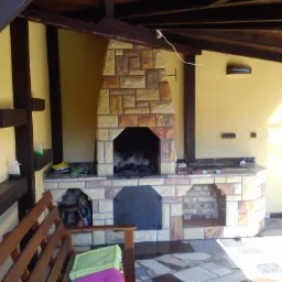 Zadaszone patio z murowanym grillem z piaskowca, drewnianymi belkami stropowymi i fragmentem drewnianego krzesła z zieloną poduszką.