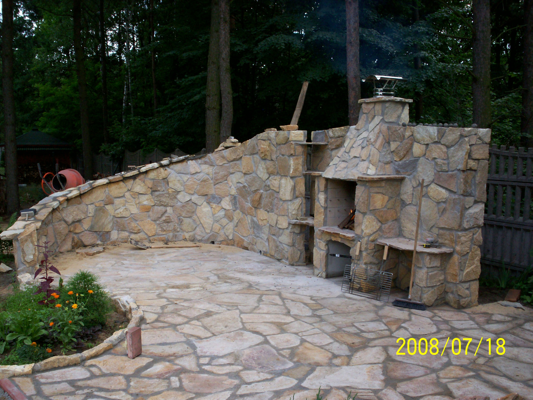 Kamienna zabudowa grilla z kominkiem i wędzarnią, wykończona nieregularną kamienną posadzką, na tle zieleni drzew.