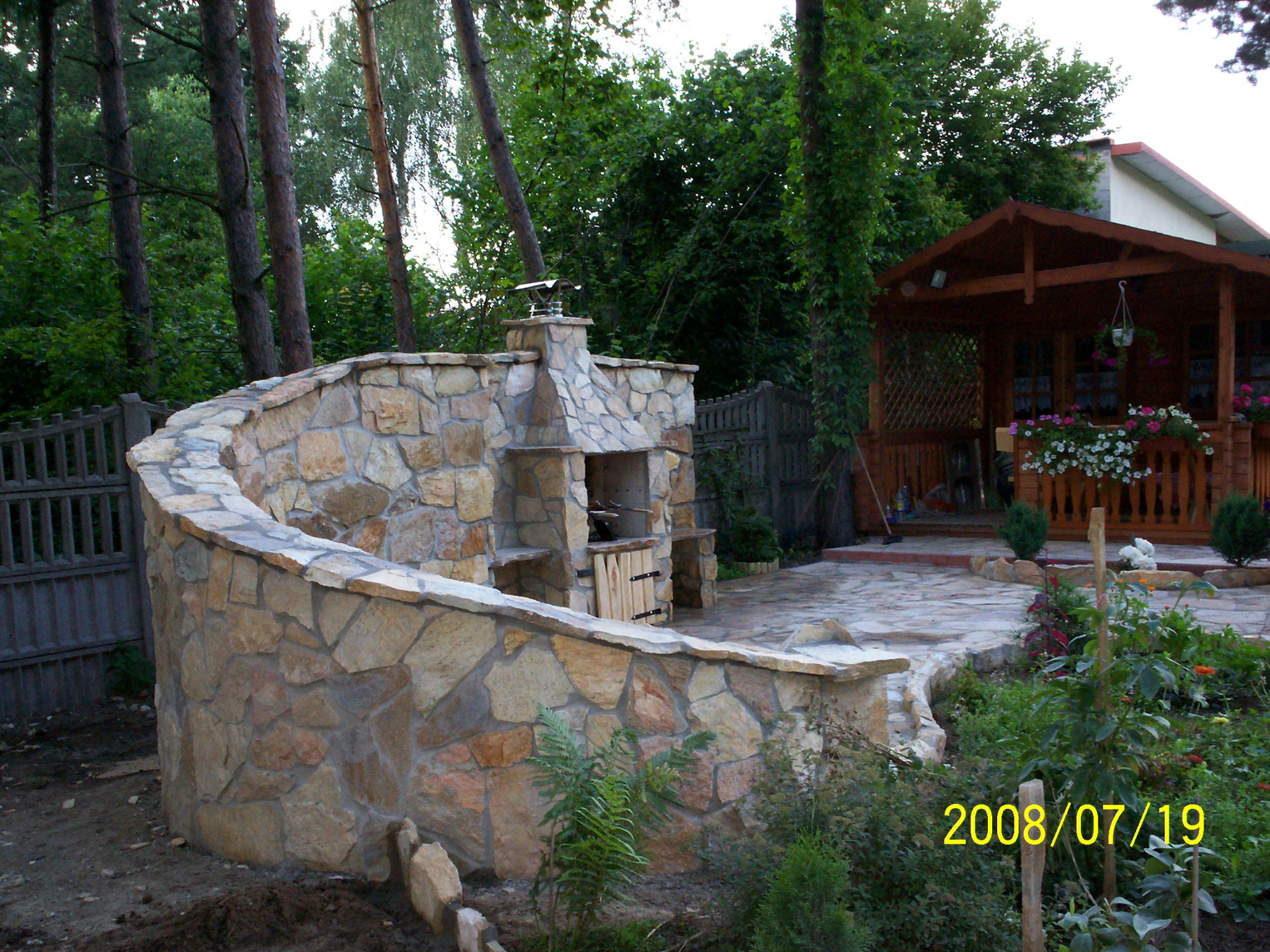 Kamienny grill ogrodowy z półkami i zamykaną szafką, otoczony wybrukowanym tarasem i zielenią, w tle drewniana altana i drzewa.