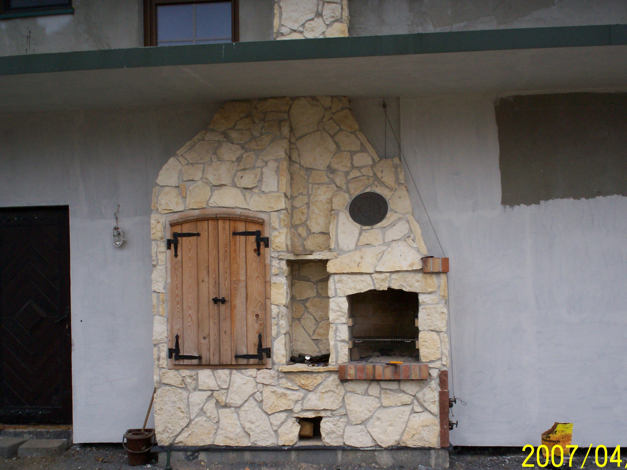 Murowany grill z kamienia z drewnianymi okiennicami, obok ciemne drzwi, widoczne ślady niedawnych prac budowlanych na ścianie.