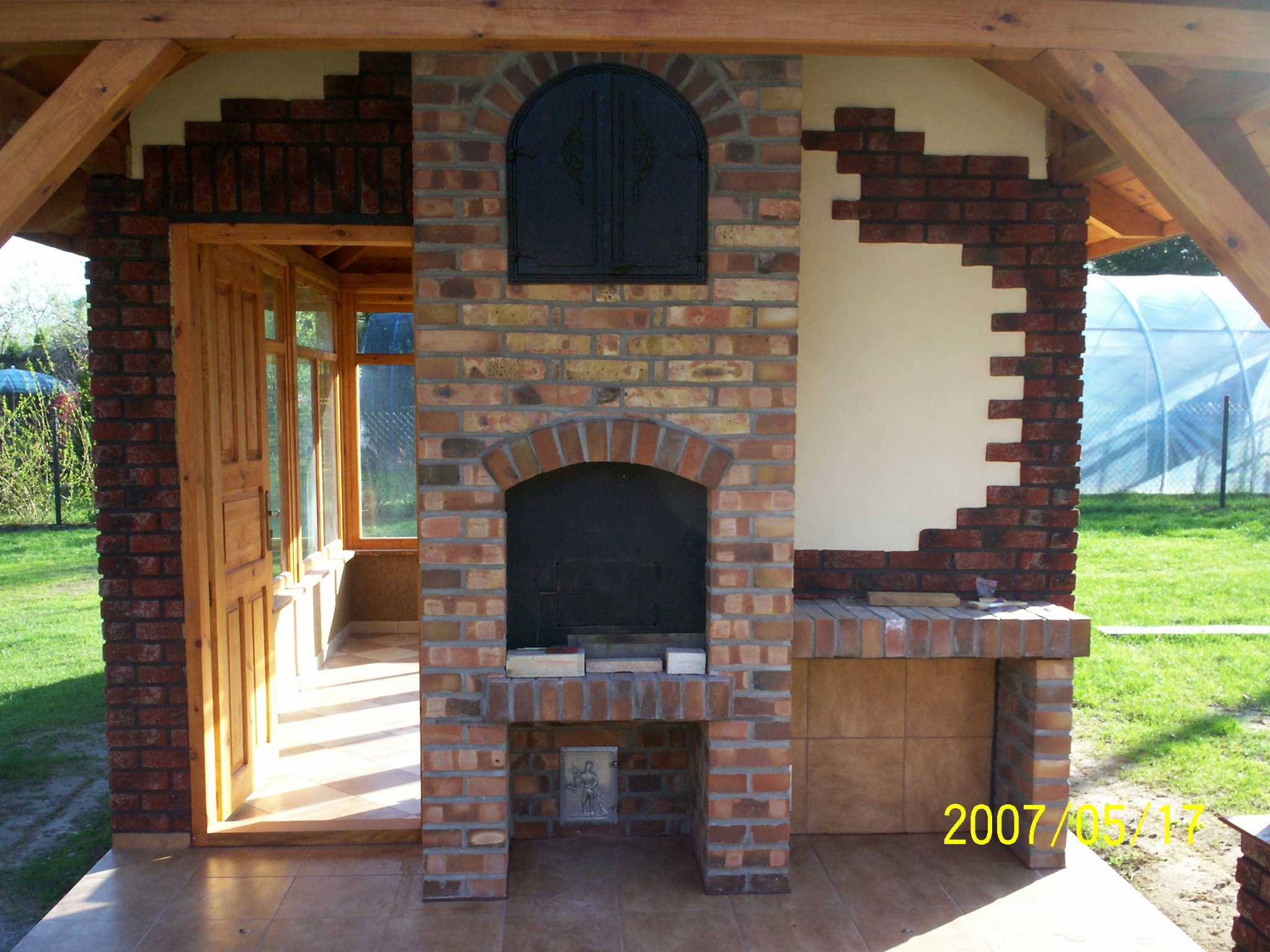 Murowany grill zewnętrzny z cegły klinkierowej, z ozdobnym zamknięciem paleniska i wnęką na akcesoria, zadaszony drewnianą konstrukcją, z widoczną datą wykonania.