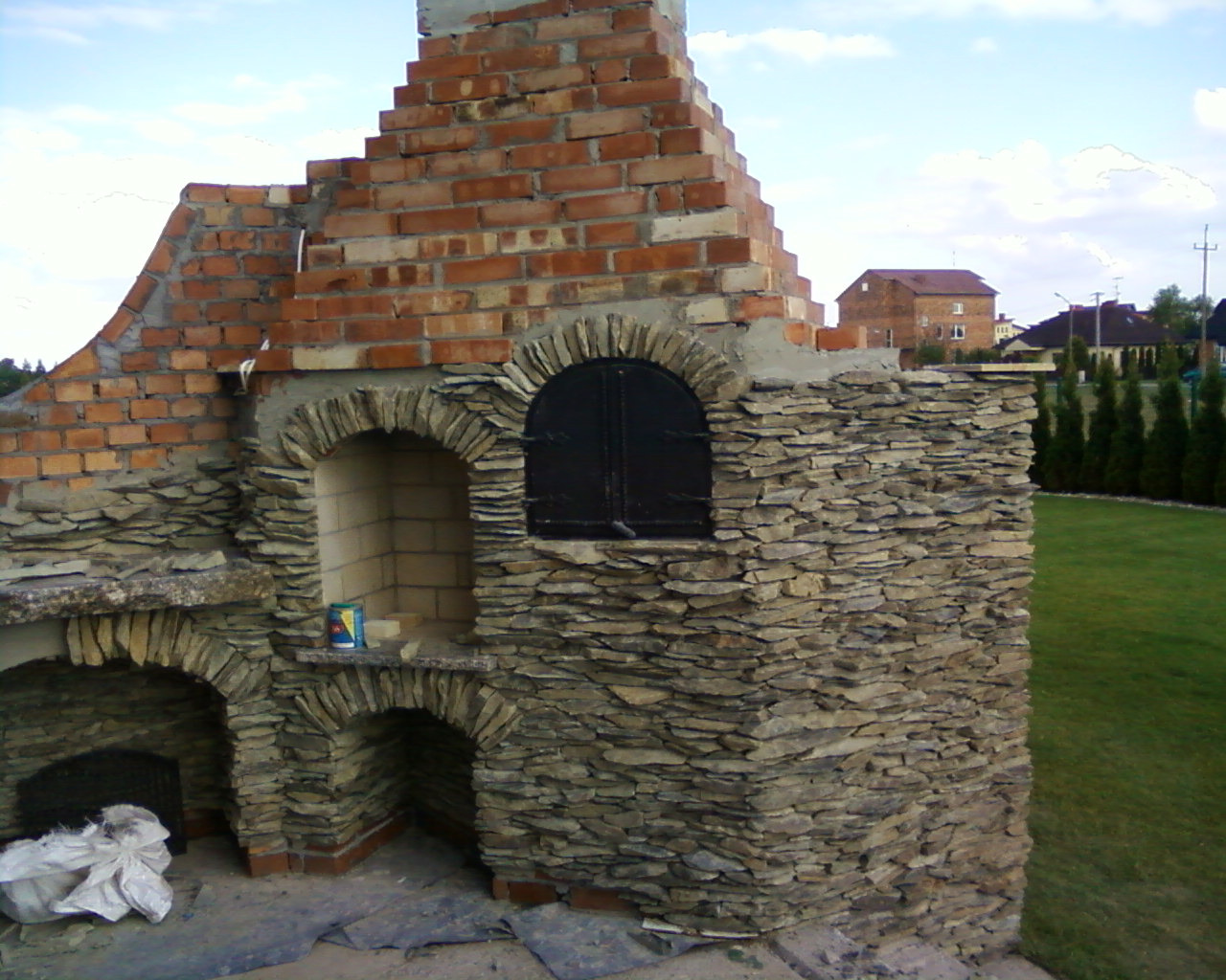 Murowany grill ogrodowy w trakcie budowy, z elementami z cegły i kamienia łupanego, widoczny niedokończony komin i czarne drzwiczki do wędzarni.