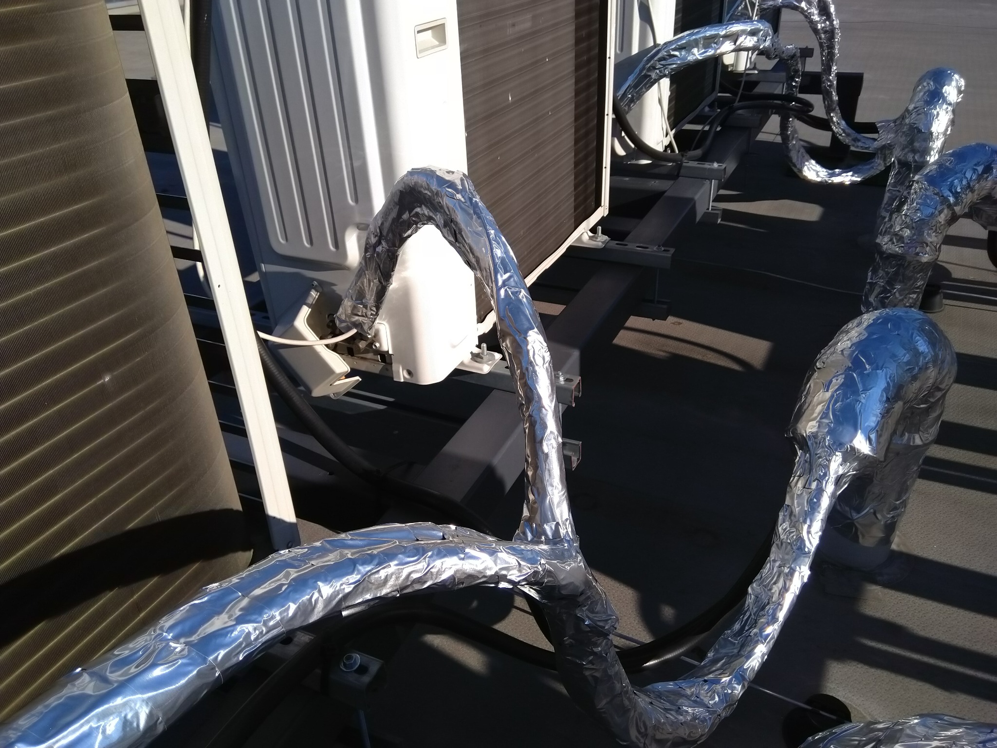 Instalacja klimatyzacji na dachu budynku, rury owinięte srebrną folią izolacyjną, widoczne jednostki zewnętrzne klimatyzatorów.