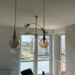 Nowoczesna lampa sufitowa z sześcioma szklanymi kloszami w kolorze bursztynowym, zawieszona w jasnym pomieszczeniu z oknami wychodzącymi na osiedle mieszkaniowe; widoczna ściana z tapetą...
