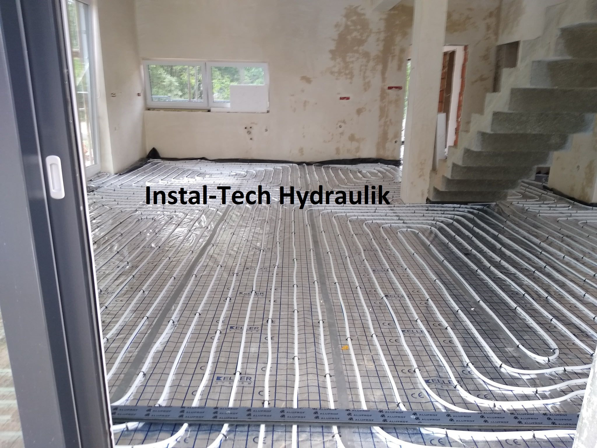 Białe rury ogrzewania podłogowego ułożone na czarnej folii izolacyjnej w surowym wnętrzu domu w trakcie budowy, widoczne schody i okno.