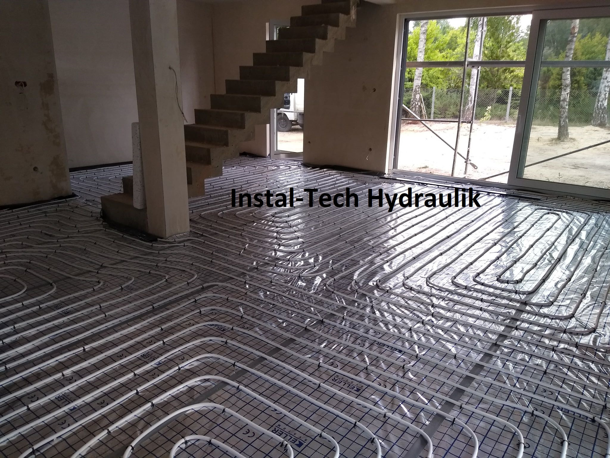 Białe rurki ogrzewania podłogowego ułożone na folii z siatką, widok na surowy beton i schody w tle, częściowo przeszklone drzwi balkonowe.