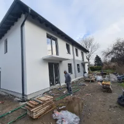 Ocieplony, pomalowany na biało dom jednorodzinny w trakcie prac wykończeniowych wokół budynku. Widoczny pracownik w kombinezonie ochronnym oraz materiały budowlane na posesji.