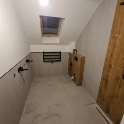 Wnętrze łazienki w trakcie remontu z widoczną instalacją podtynkową, grzejnikiem drabinkowym i drewnopodobną obudową WC, wykończone szarą płytką imitującą marmur, z oknem dachowym.