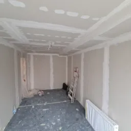 Pokój w trakcie prac wykończeniowych: ściany obłożone płytami kartonowo-gipsowymi, widoczne łączenia szpachlowane, sufit z dekoracyjnymi elementami z płyt gipsowych, drabina oparta o ścianę...