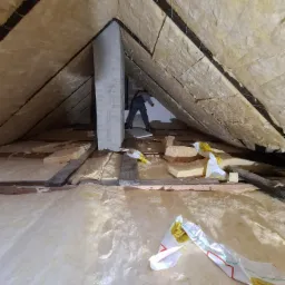 Prace izolacyjne poddasza z widocznym pracownikiem montującym wełnę mineralną między krokwiami, fragmenty opakowań izolacji na podłodze