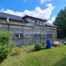 Dom jednorodzinny w trakcie tynkowania elewacji, widoczne rusztowanie, pracownik na rusztowaniu i wiadro z zaprawą na dachu, błękitne niebo z chmurami w tle.