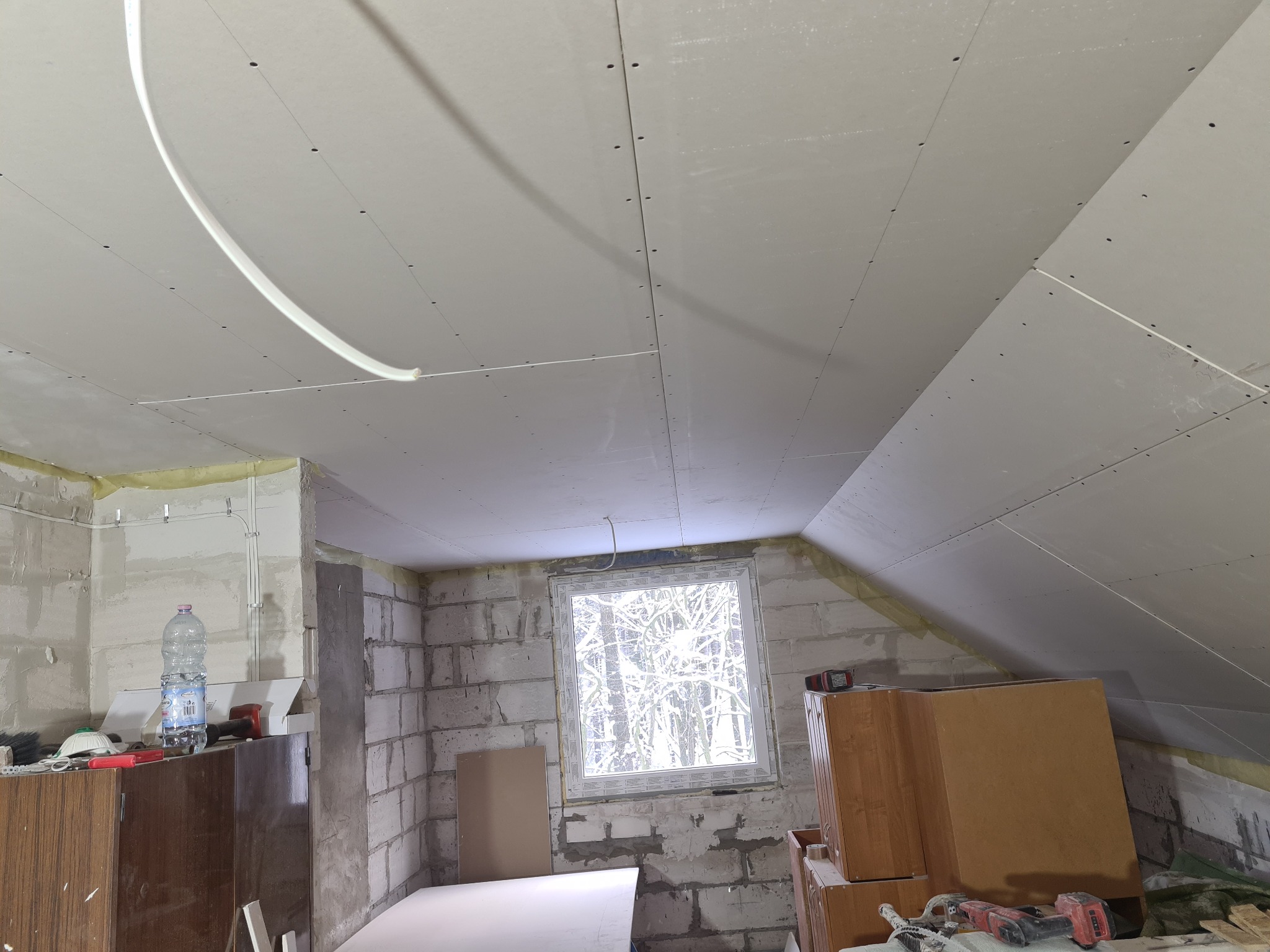 Poddasze w trakcie prac wykończeniowych: zamontowane płyty kartonowo-gipsowe na skośnym suficie, widoczne przewody elektryczne, ściany z surowych bloczków, okno z widokiem na ośnieżone drzewa...