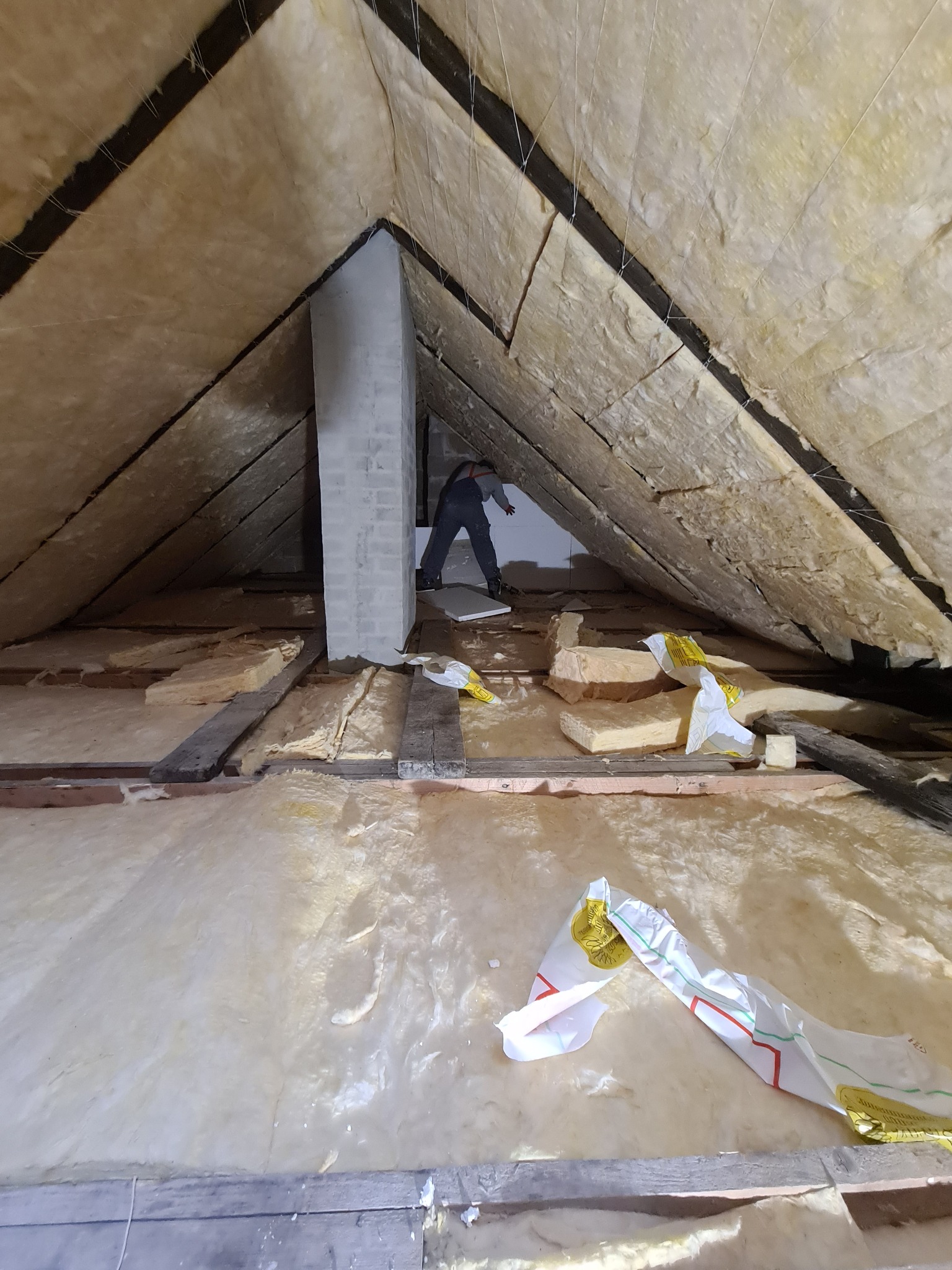 Prace izolacyjne poddasza z widocznym pracownikiem montującym wełnę mineralną między krokwiami, fragmenty opakowań izolacji na podłodze