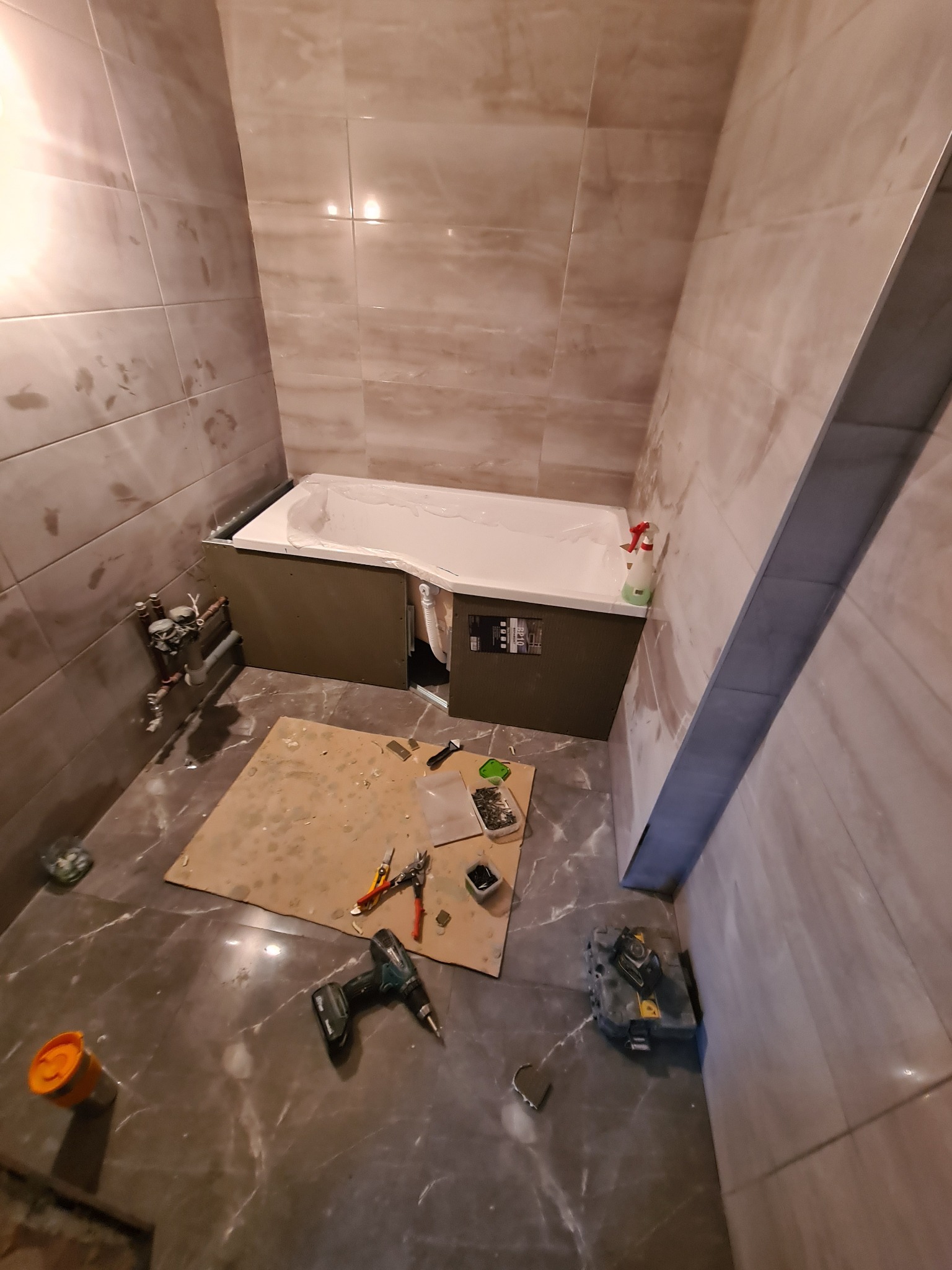 Montaż obudowy wanny w łazience wyłożonej marmurkowymi płytkami, widoczne narzędzia pracy: wiertarka, kombinerki, nożyk na kartonie, butelka z płynem czyszczącym obok wanny.