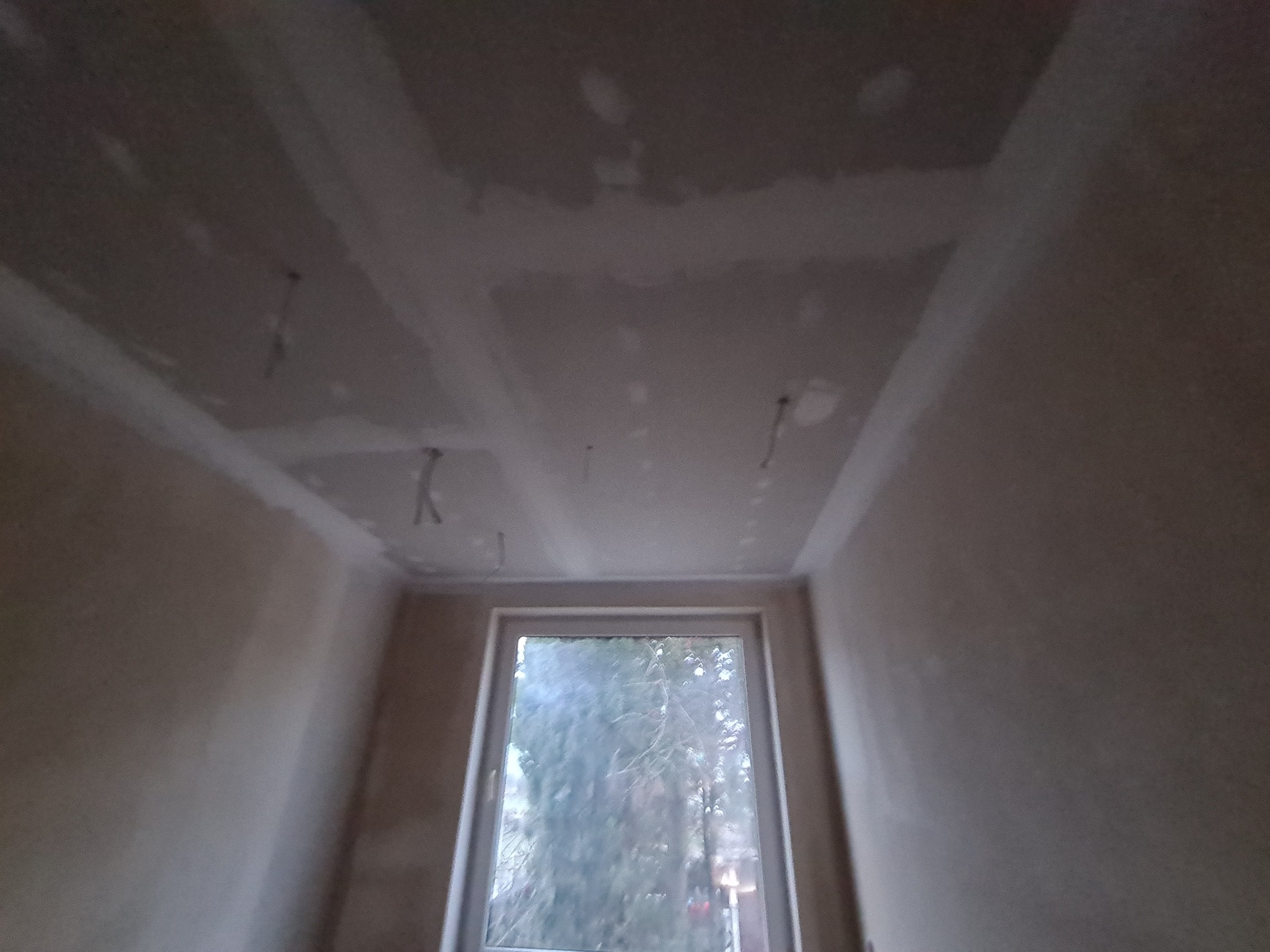 Pokój w trakcie remontu: sufit i ściany po nałożeniu gładzi szpachlowej, widoczne wystające kable elektryczne, okno z widokiem na zewnątrz.