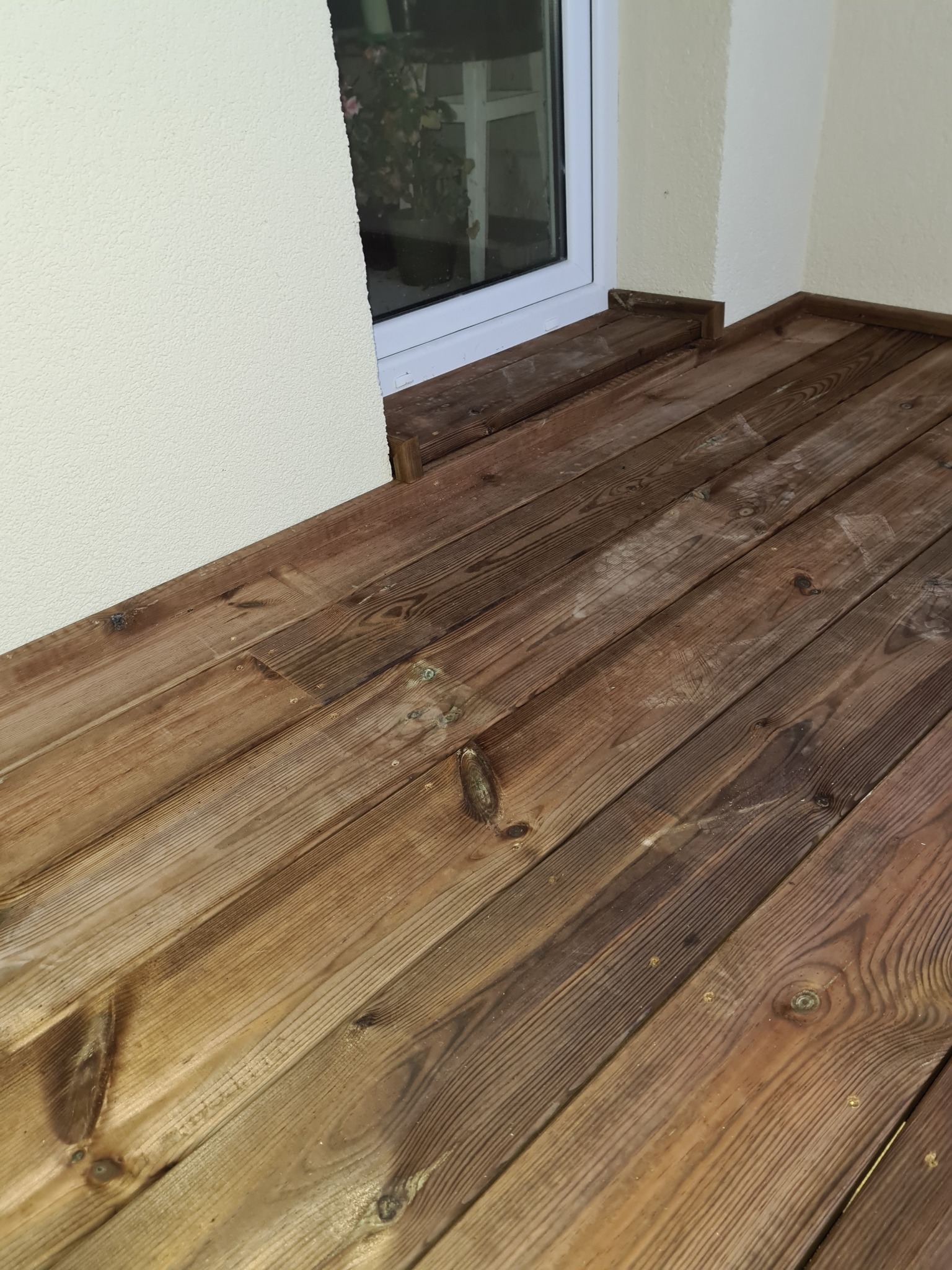Drewniana podłoga balkonu z widocznymi śladami użytkowania, przy białym oknie i kremowej ścianie.