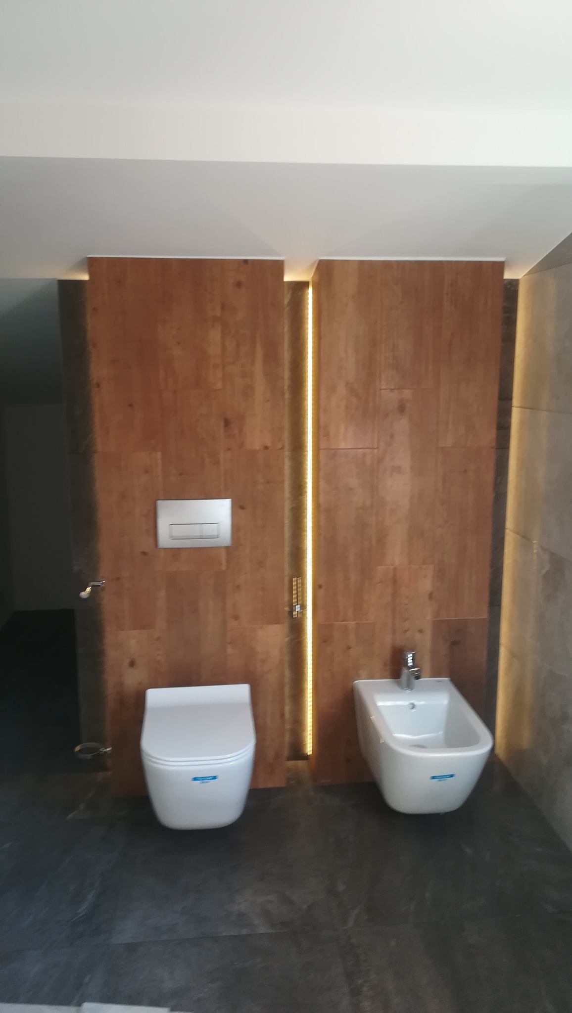 Nowoczesna łazienka z drewnianą okładziną ściany, podświetleniem LED, wiszącą toaletą i bidetem na tle ciemnej podłogi.