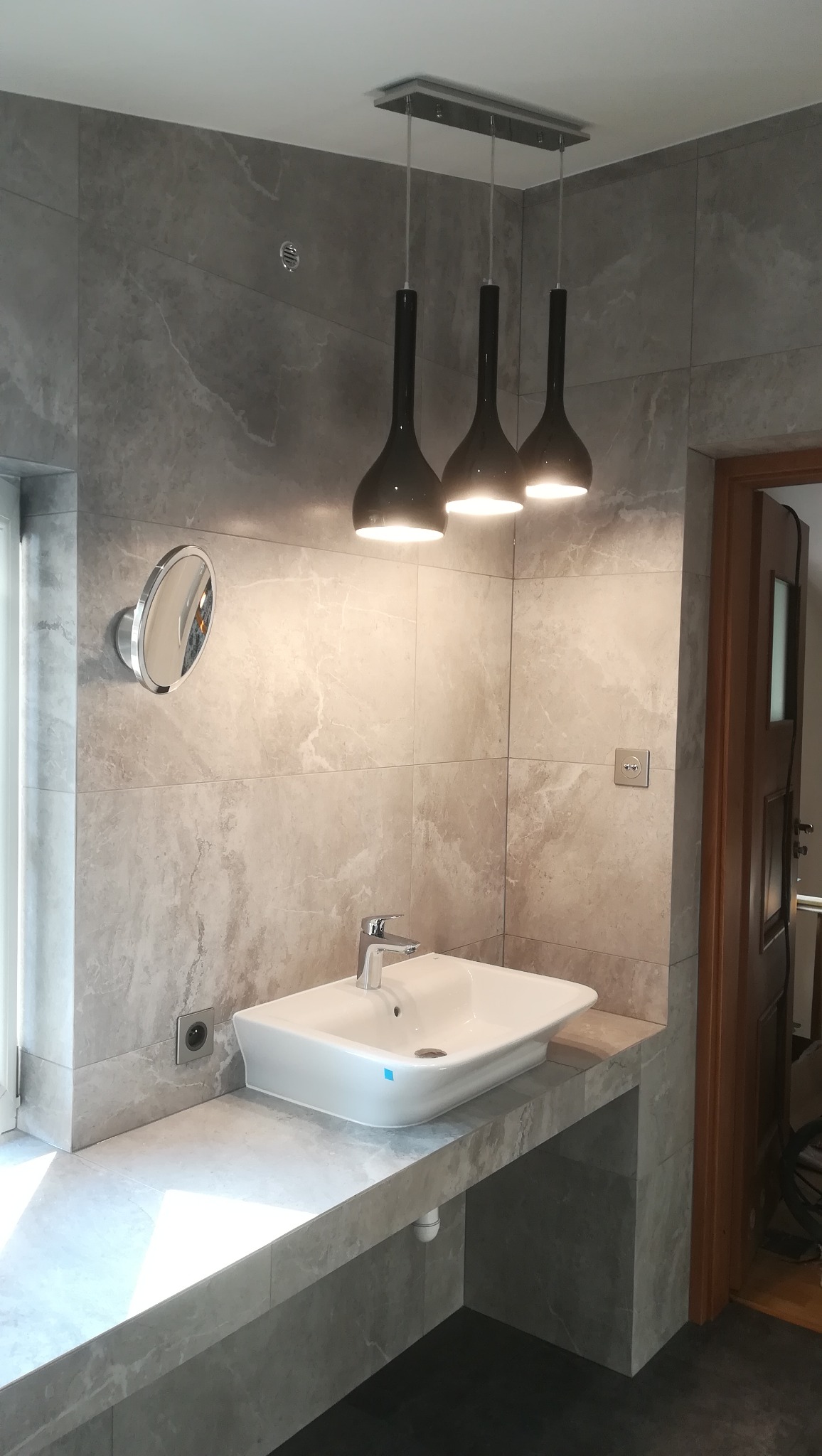 Nowoczesna łazienka z szarymi płytkami imitującymi marmur, białą umywalką nablatową, chromowaną baterią i okrągłym lustrem. Nad umywalką wiszą trzy czarne, podłużne lampy. Widoczny fragment...