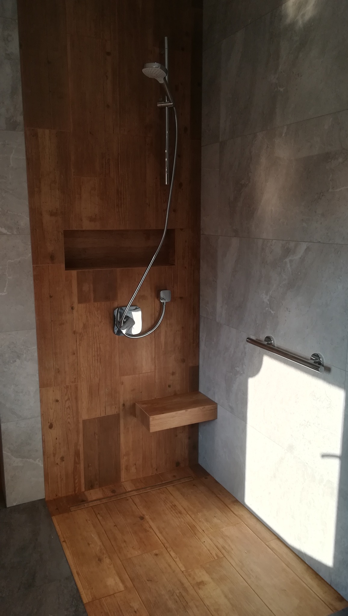 Nowoczesna kabina prysznicowa z drewnianymi panelami ściennymi i podłogą, z wnęką i siedziskiem z drewna, oraz chromowaną armaturą i uchwytem dla niepełnosprawnych.