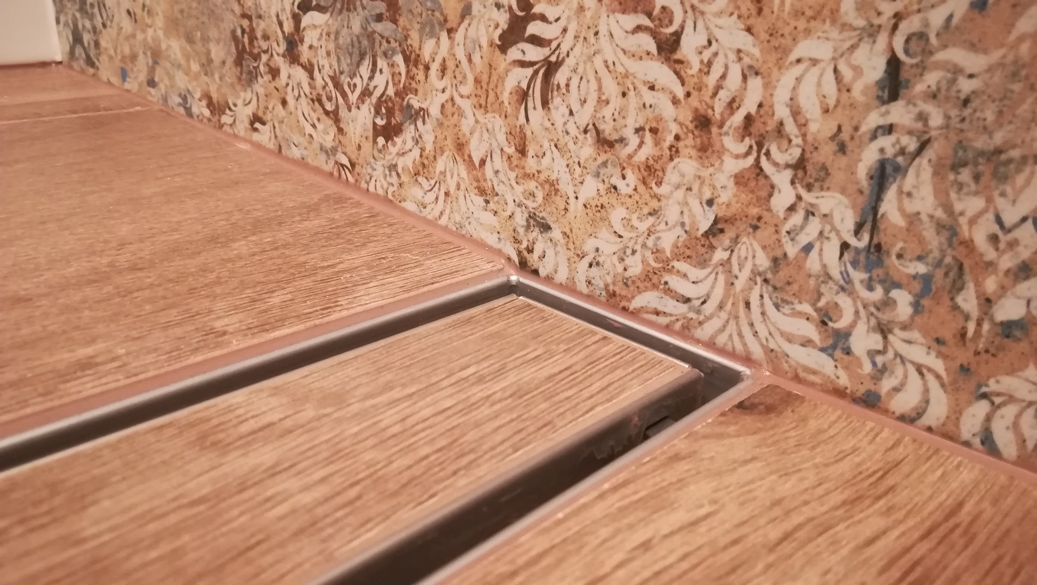 Ułożone płytki podłogowe imitujące drewno z odpływem liniowym w kabinie prysznicowej, wzorzyste płytki ścienne w tle.