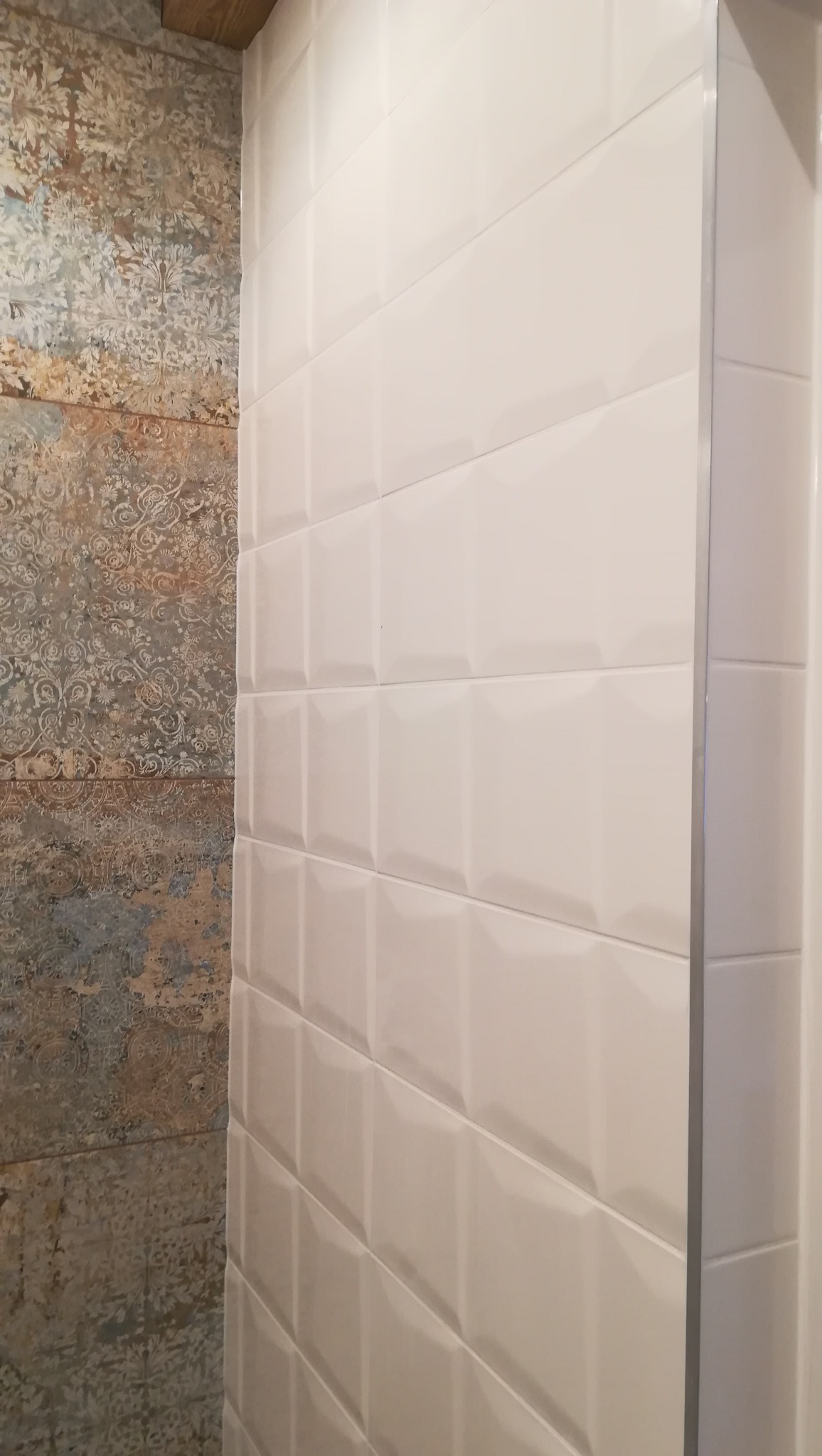 Ściana wykończona białymi, wypukłymi płytkami ceramicznymi ułożonymi poziomo, sąsiadująca z ścianą o wzorzystej, postarzanej fakturze w odcieniach beżu i błękitu, widoczne z bliska.