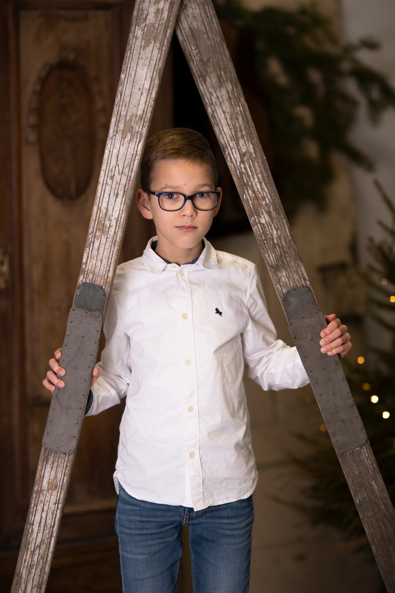 Portret chłopca w okularach, opierającego się o starą, drewnianą drabinę na tle świątecznym.