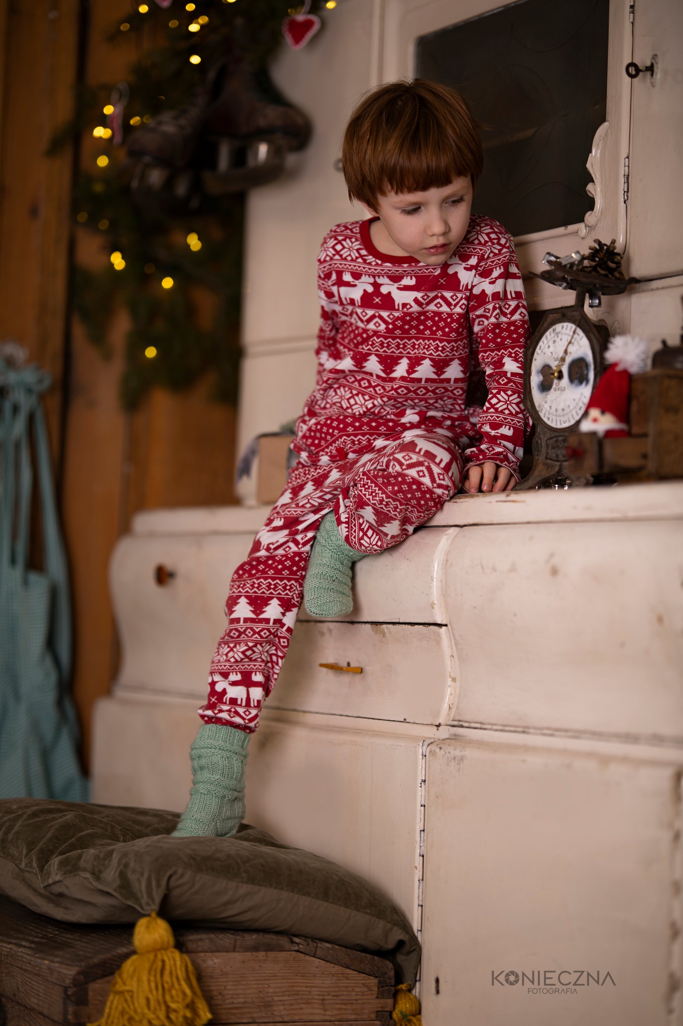 Chłopiec w świątecznej piżamie siedzi na starym kredensie, obok waga i dekoracje świąteczne, w tle choinka z lampkami i łyżwami.