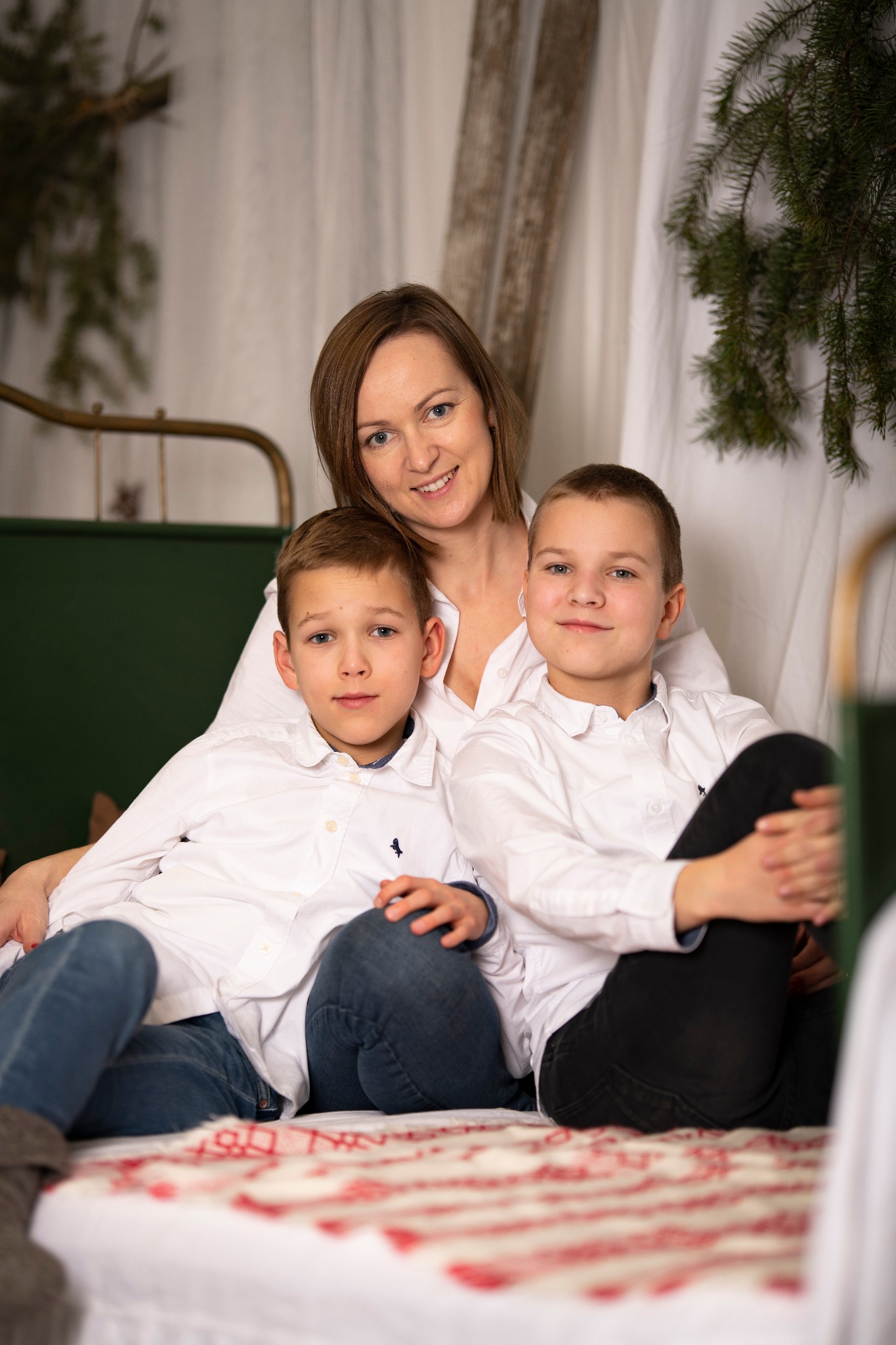 Portret rodzinny: matka z dwoma synami w białych koszulach i jeansach, na tle białej zasłony i choinki, ciepłe oświetlenie.