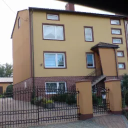 Dwupiętrowy dom z żółtą elewacją i ceglaną podmurówką, widoczny zza czarnego, kutego ogrodzenia. Charakterystyczny daszek nad wejściem z zewnętrznymi schodami. Dom po przebudowie.