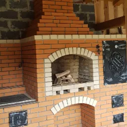 Murowany z cegły kominek zewnętrzny z paleniskiem, rusztem i zapasem drewna, oświetlony wiszącą lampą, z elementami dekoracyjnymi z kutego żelaza.