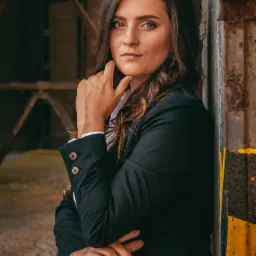 Portret kobiety w czarnej marynarce opierającej się o metalową ścianę z żółto-czarnymi pasami, w tle widoczna konstrukcja.