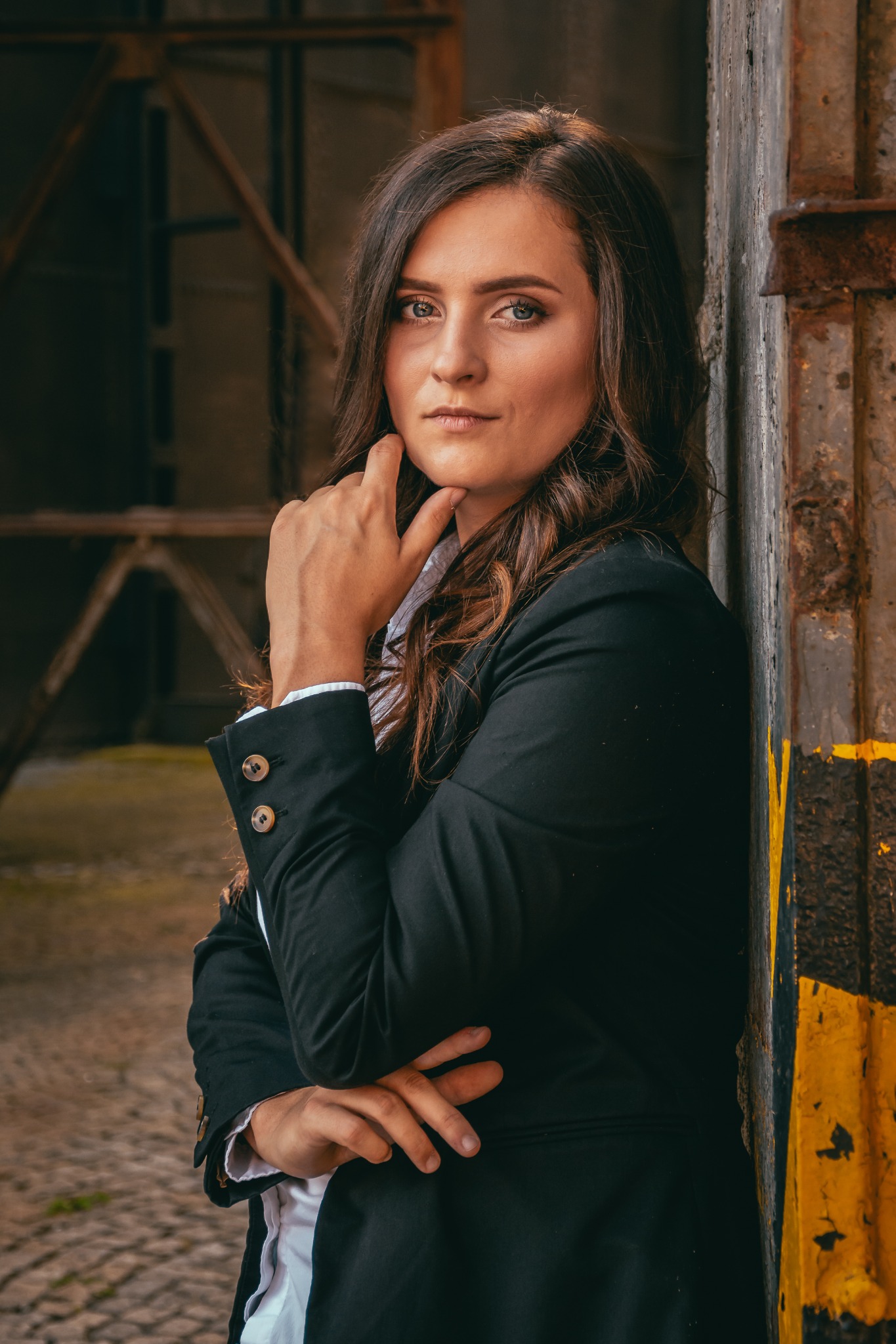 Portret kobiety w czarnej marynarce opierającej się o metalową ścianę z żółto-czarnymi pasami, w tle widoczna konstrukcja.