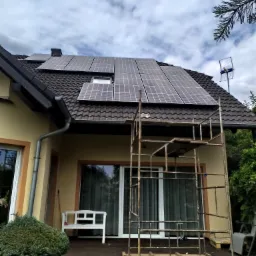 Montaż paneli słonecznych na dachu domu jednorodzinnego, widoczne rusztowanie i antena telewizyjna.