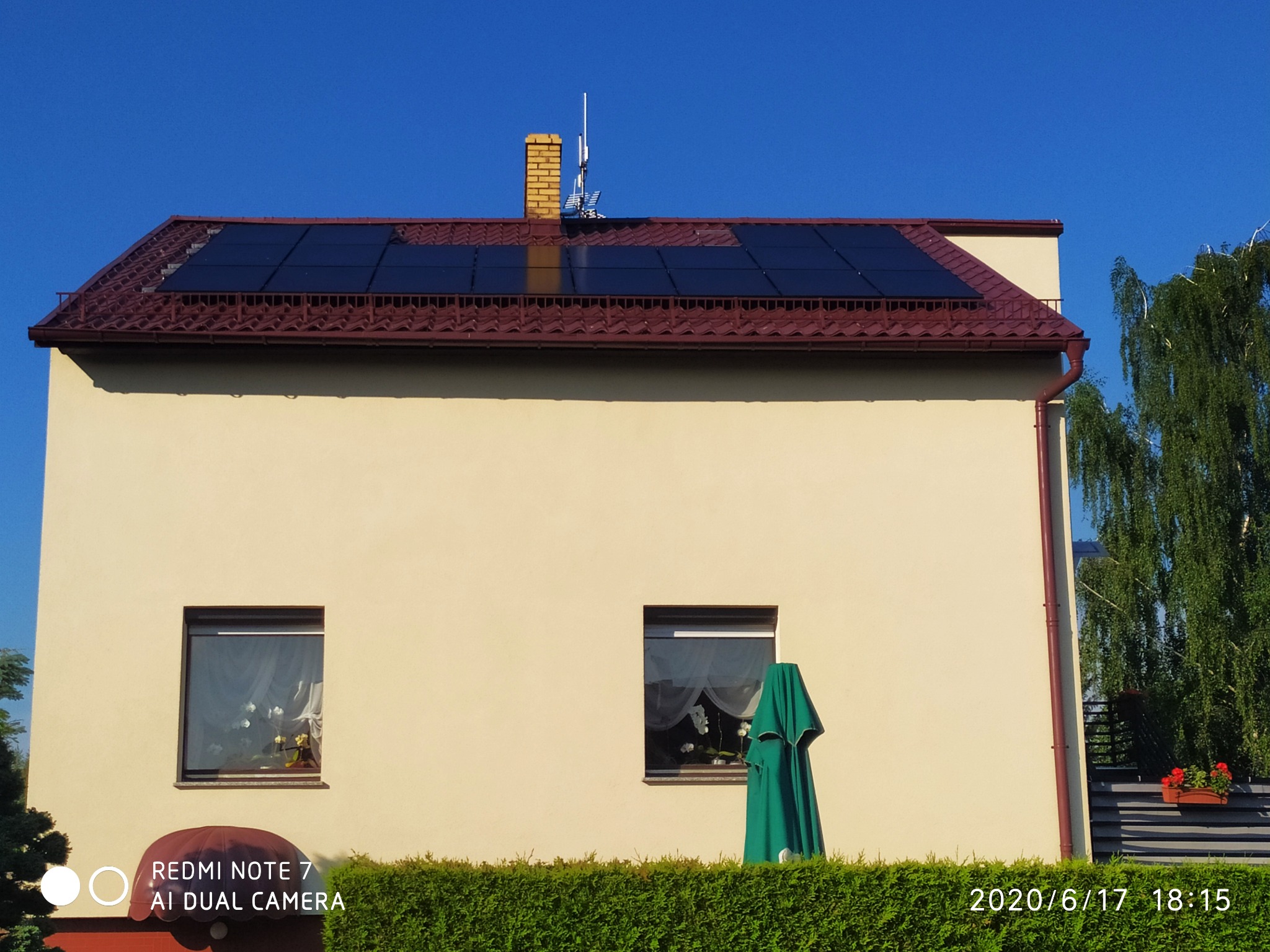 Dwukondygnacyjny dom jednorodzinny z zainstalowanymi panelami słonecznymi na dachu z czerwonej dachówki, widoczny komin z anteną oraz starannie przycięty żywopłot przed budynkiem. Częściowo...