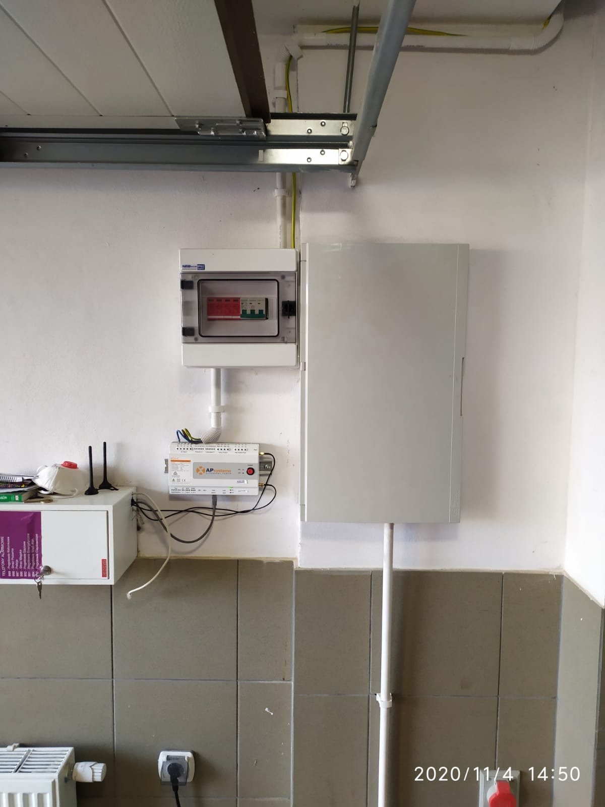 Szara skrzynka elektryczna z bezpiecznikami i aparaturą sterującą zamontowana na ścianie z szarymi płytkami, widoczne przewody i elementy instalacji elektrycznej.
