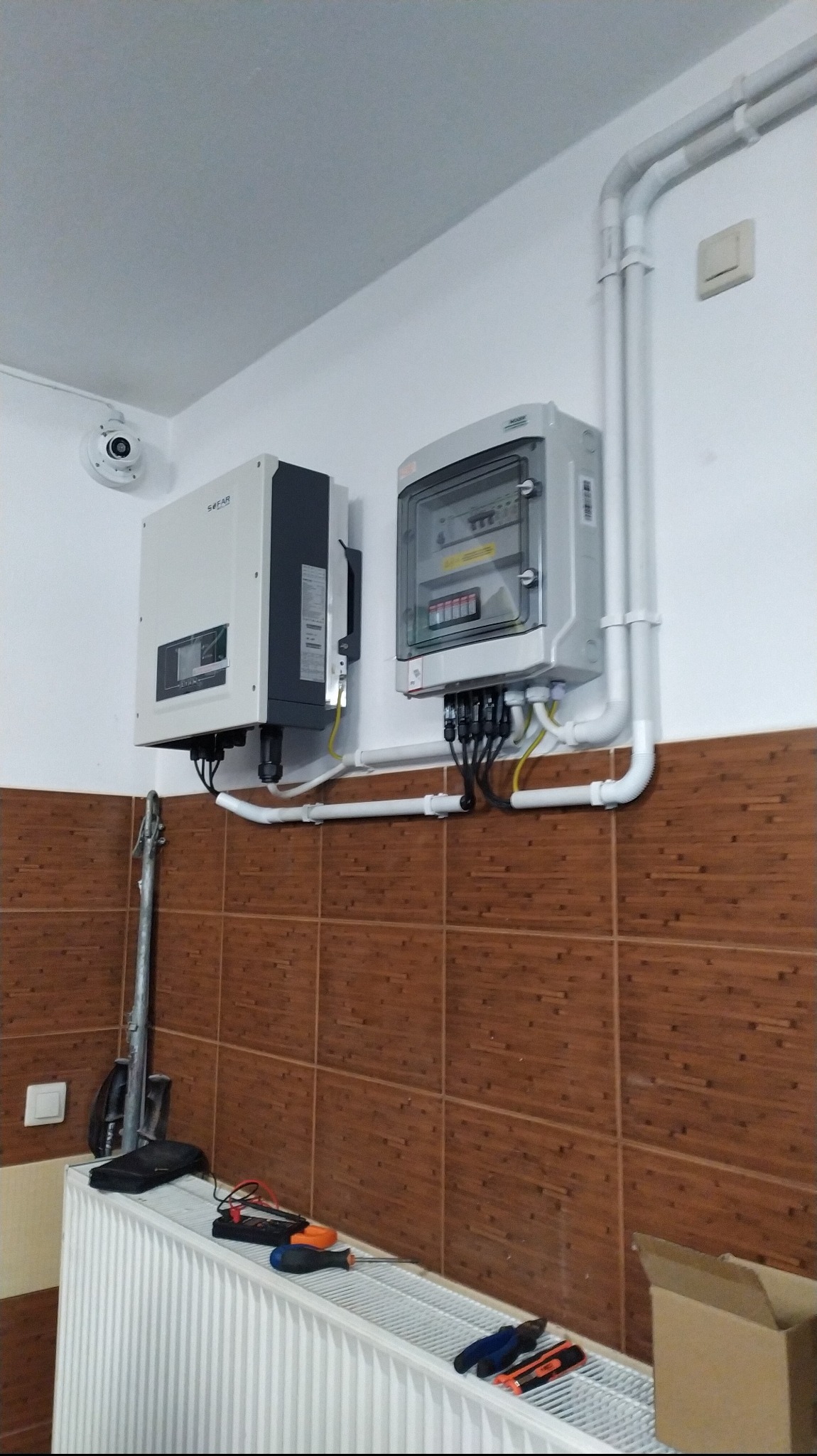Zamontowany inwerter fotowoltaiczny marki Sofar Solar i skrzynka zabezpieczeń na ścianie z brązowymi płytkami, widoczne przewody, rury PCV, kamera monitoringu na suficie oraz narzędzia i miernik...