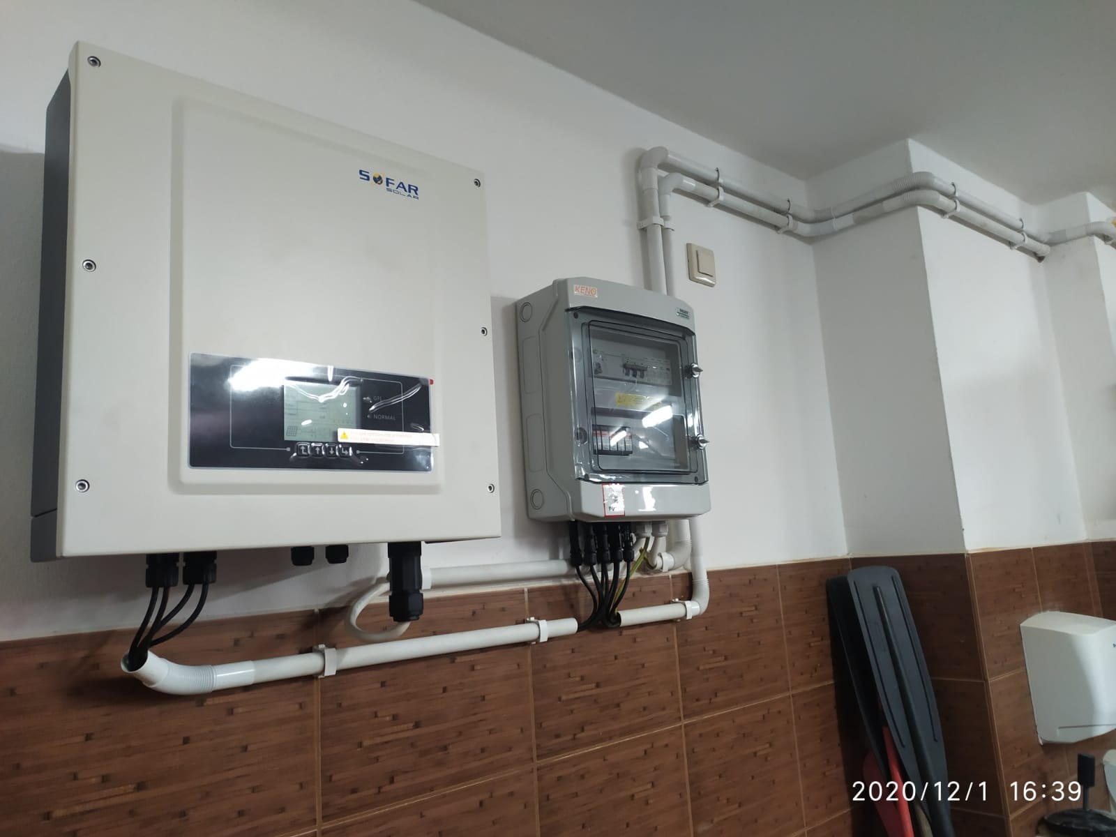 Zamontowany inwerter fotowoltaiczny firmy SOFAR oraz skrzynka zabezpieczeń na ścianie z brązowymi płytkami, widoczne przewody i rury instalacyjne.