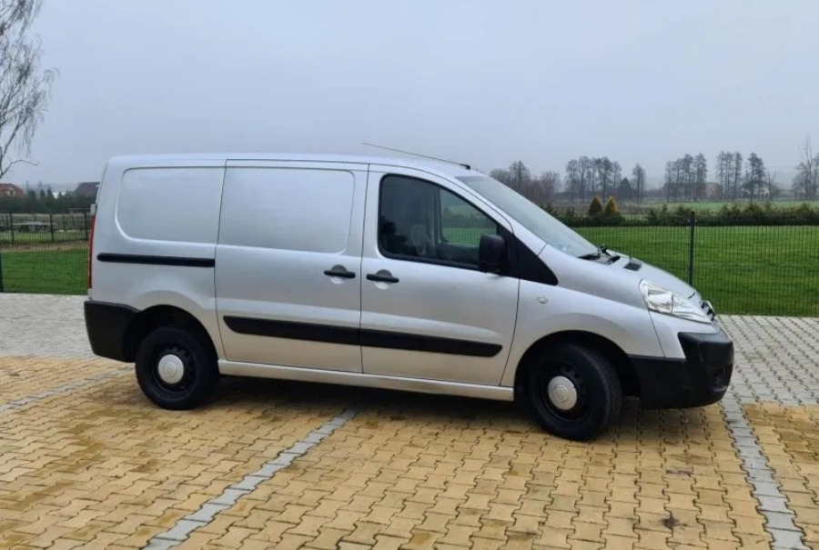 Srebrny samochód dostawczy Peugeot Expert z czarnymi kołpakami, zaparkowany na kostce brukowej w pochmurny dzień.